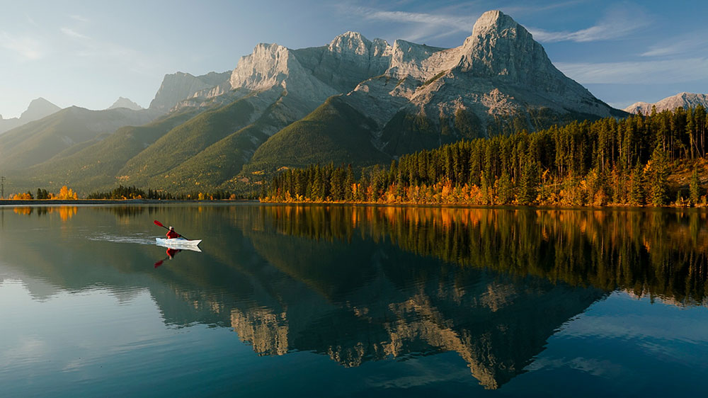 Calm mountain lake at Lake Louise, Alberta, with a single canoe on the water, evergreen forest along the shoreline, and steep rocky peaks reflected clearly in the still surface under soft daylight.