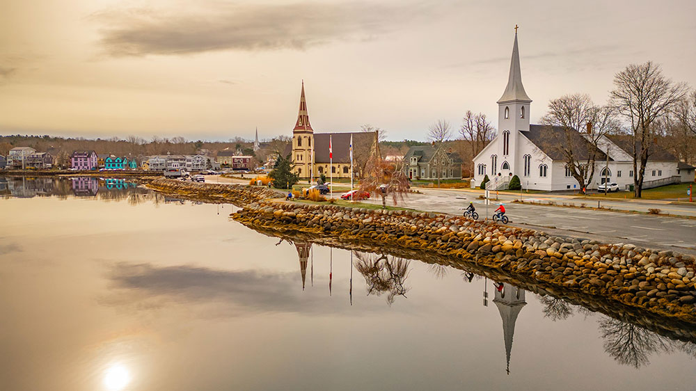 Waterfront view of Mahone Bay, Nova Scotia, with calm reflective water, the iconic three church steeples along the shoreline, colorful houses, and a stone seawall under soft evening light.