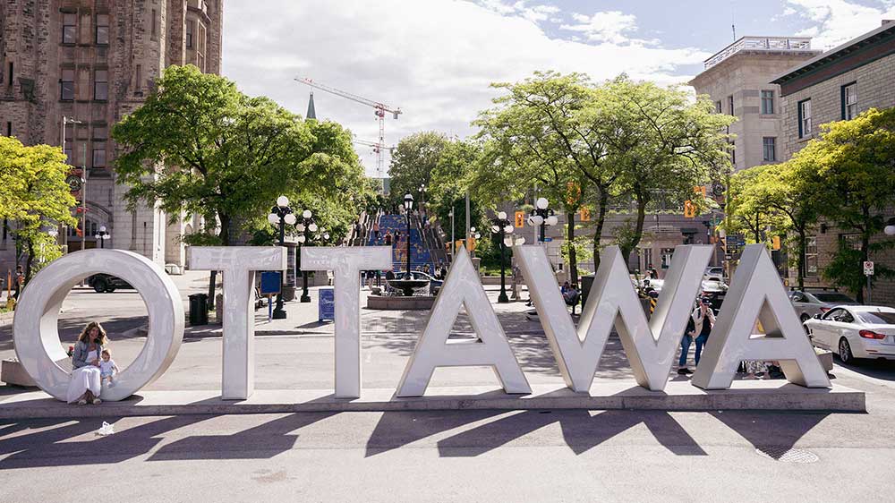 Large white sign saying "Ottawa" in capital letters in downtown Ottawa with trees, historic buildings, and the blue stairs in the background.