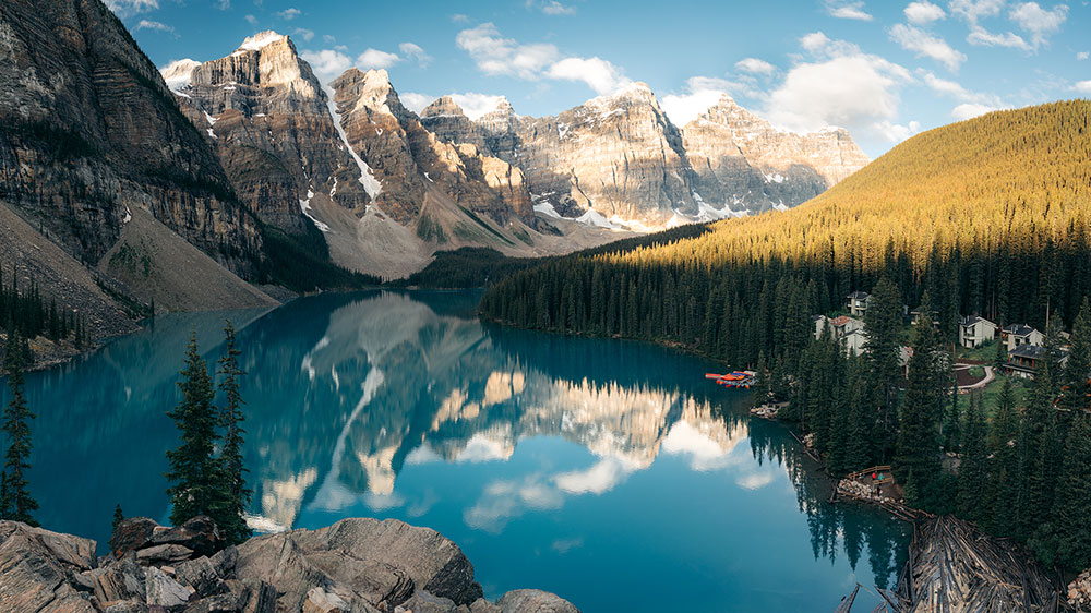Calm mountain lake at Lake Louise, Alberta, with evergreen forest along the shoreline, and steep rocky peaks reflected clearly in the still surface under soft daylight.