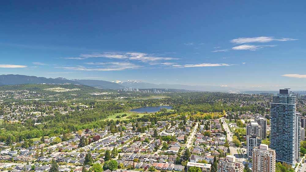 Aerial view of Burnaby, BC, with residential streets, green parkland, nearby high‑rise buildings, and mountains in the distance.