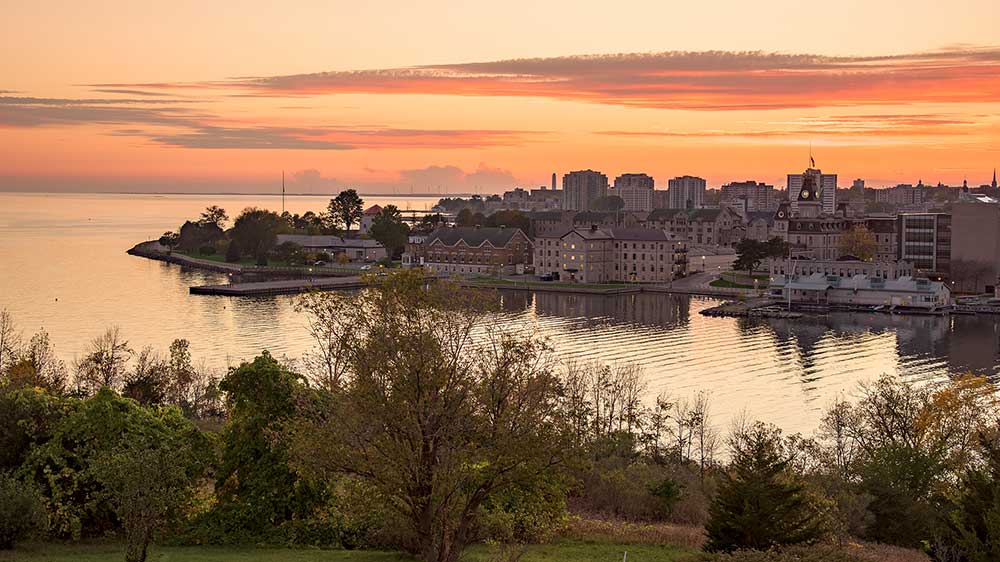 Sunset view overlooking Kingston, Ontario, with historic stone buildings and waterfront in the midground, calm water reflecting warm sky colours, and trees and grassy parkland in the foreground.