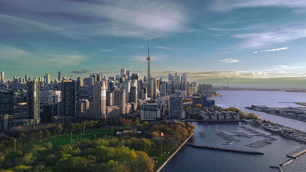 Aerial view of downtown Toronto, Ontario, with the CN Tower centered among dense high‑rise buildings, a marina and waterfront park in the foreground, and Lake Ontario extending into the distance beneath a partly cloudy sky.