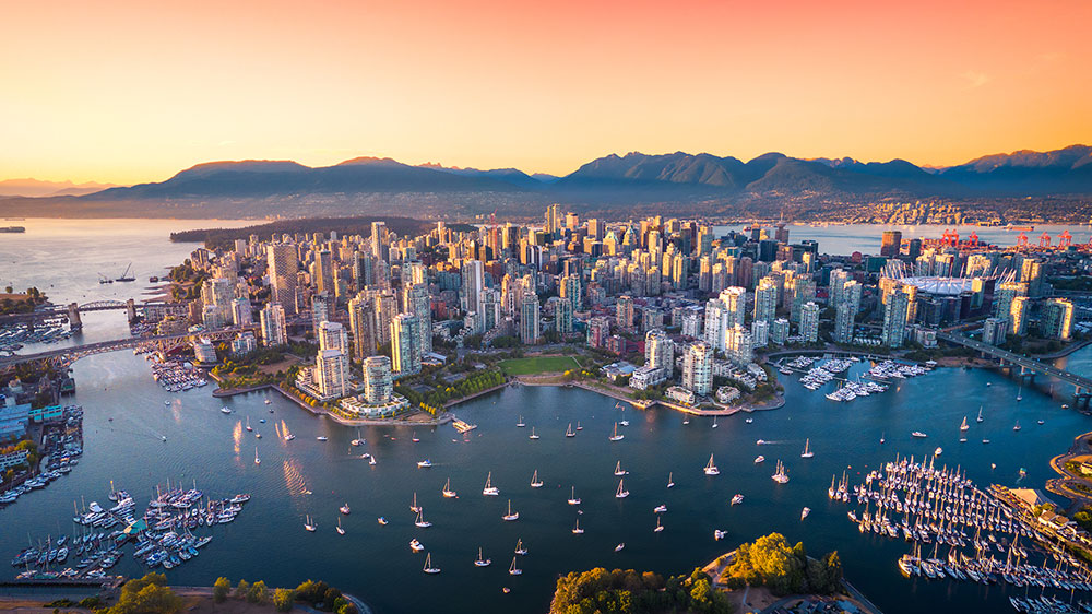Aerial view of downtown Vancouver at sunset, showing high‑rise buildings, marinas with sailboats, False Creek waterways, and mountains in the background.