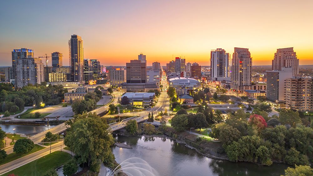 Aerial view of downtown London, Ontario, at sunset, featuring high‑rise buildings, tree‑lined streets, a river and parkland in the foreground, and city lights beginning to glow under a warm evening sky.