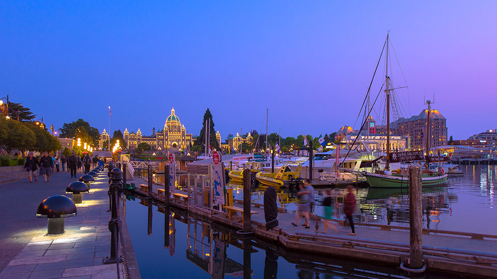 Evening view of Victoria’s Inner Harbour in British Columbia, showing docked boats, a waterfront boardwalk with pedestrians, and the brightly illuminated Parliament Buildings reflected in the harbour at blue hour.