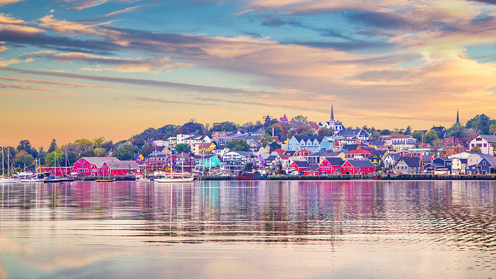 Colourful waterfront town of Lunenburg, Nova Scotia, with historic wooden buildings along the harbour, boats moored in the calm water, and soft pastel clouds reflected across the shoreline at sunset.