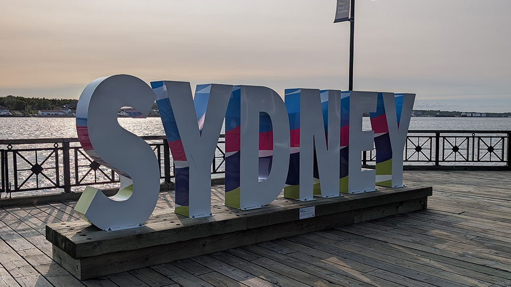 Large white sign saying "SYDNEY" in capital letters on a wooden waterfront boardwalk in Sydney, Nova Scotia, with open water behind the letters and soft evening light across the sky and harbour.