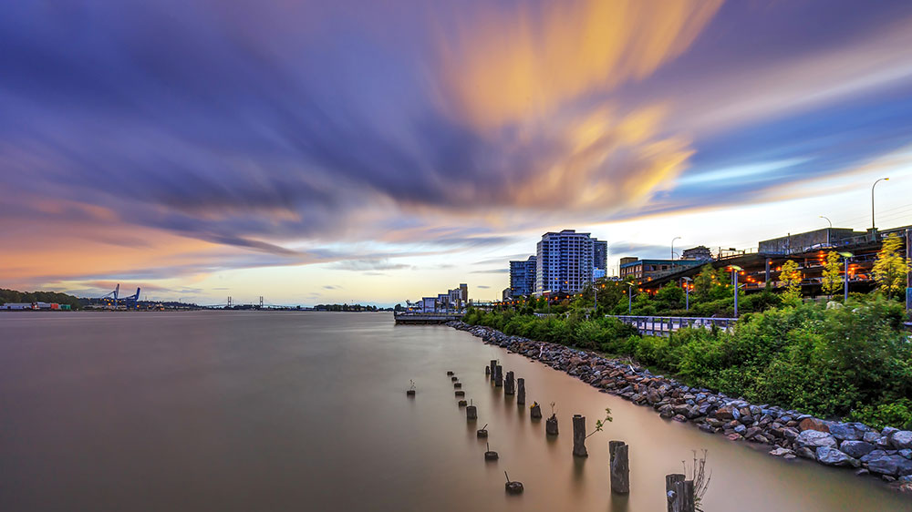 Long‑exposure view of a riverfront in New Westminster, British Columbia, featuring calm water with wooden pilings in the foreground, a lit waterfront walkway and buildings along the shore, and dramatic streaked clouds across the evening sky.